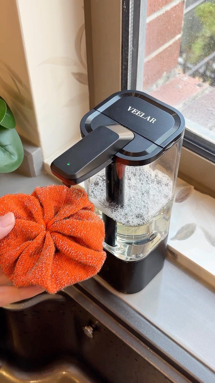 Automatic soap dispenser dispensing liquid onto an orange scrubbing sponge by a window.
