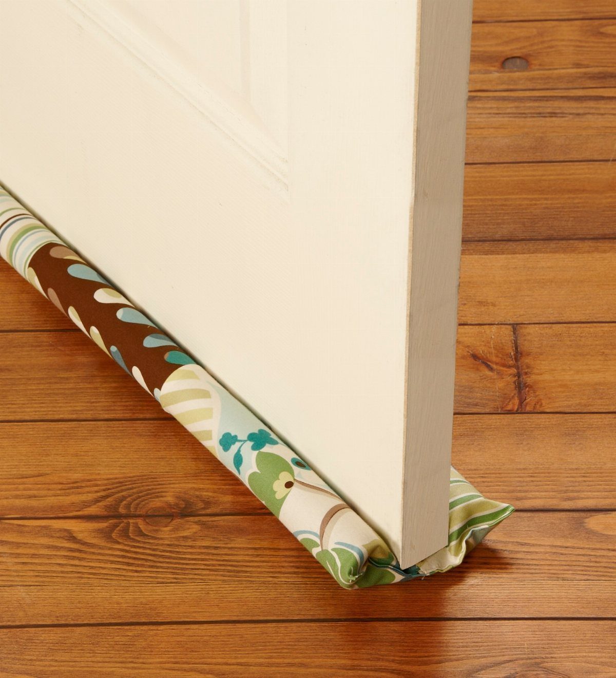 Floral fabric draft stopper blocking the gap under a white door on wood flooring.