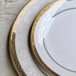 White china plates with ornate gold rims and embossed floral patterns on grey background