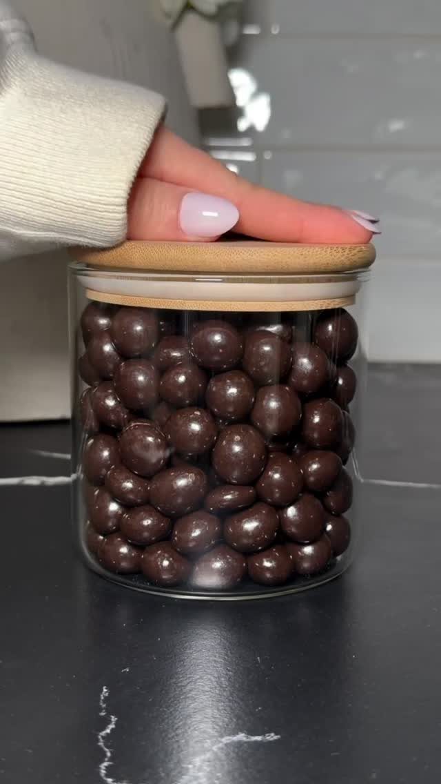 17 Kitchen Snack Storage Ideas for Pantries and Counters 18 Glass jar with wooden lid filled with chocolate snacks on a dark counter.