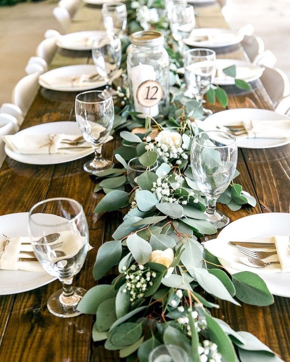 26 Table Toppers Wedding Ideas, From Candlelit to Tropical 4 Green eucalyptus garland with white flowers as a centerpiece on a wooden wedding table.