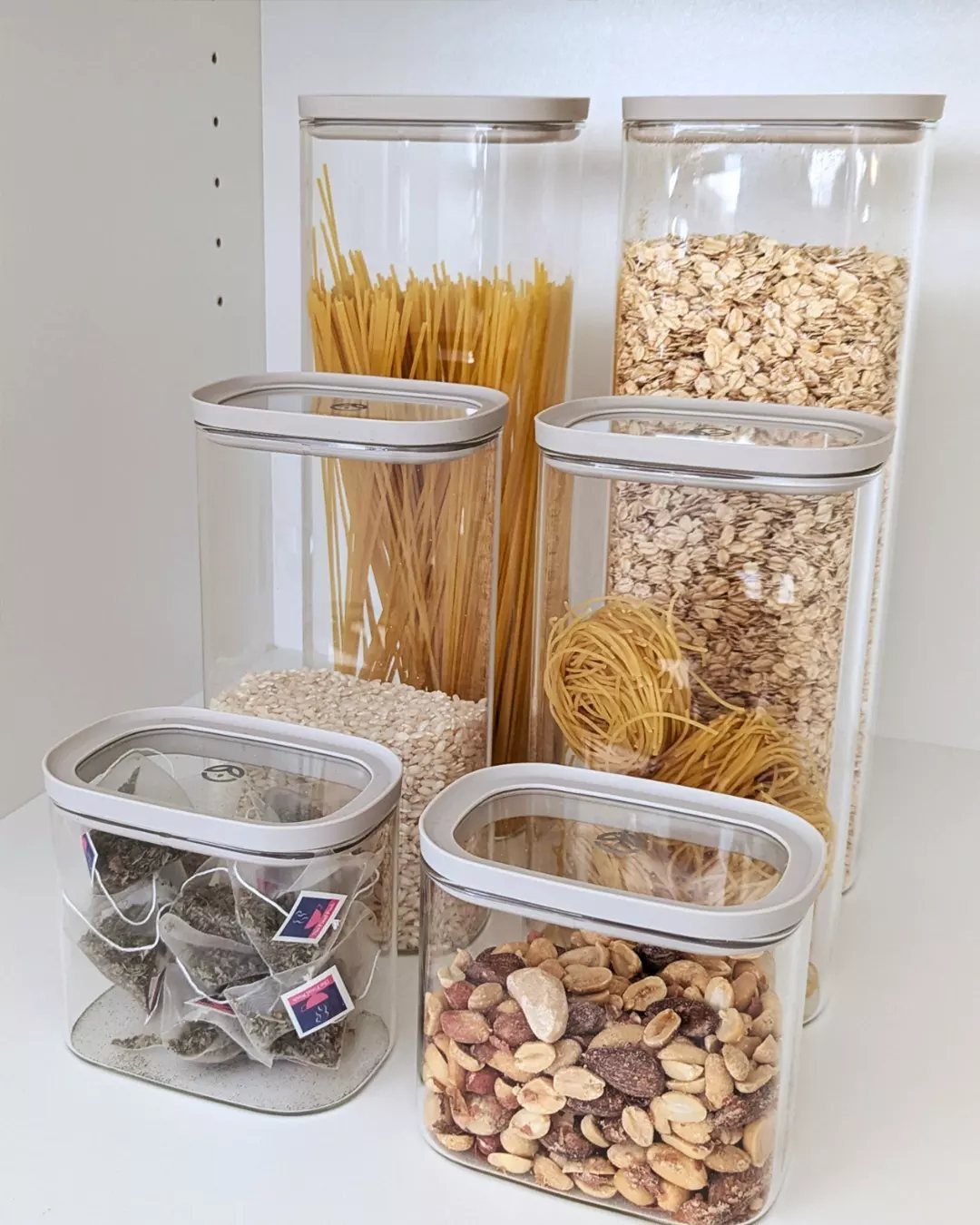 17 Kitchen Snack Storage Ideas for Pantries and Counters 5 Matching glass canisters filled with pasta, oats, and nuts on a white shelf.