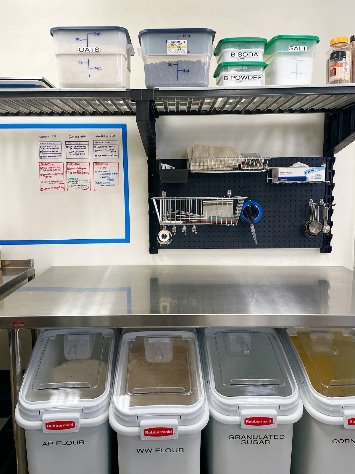 Stainless steel kitchen counter with black metal shelves and large ingredient storage bins.