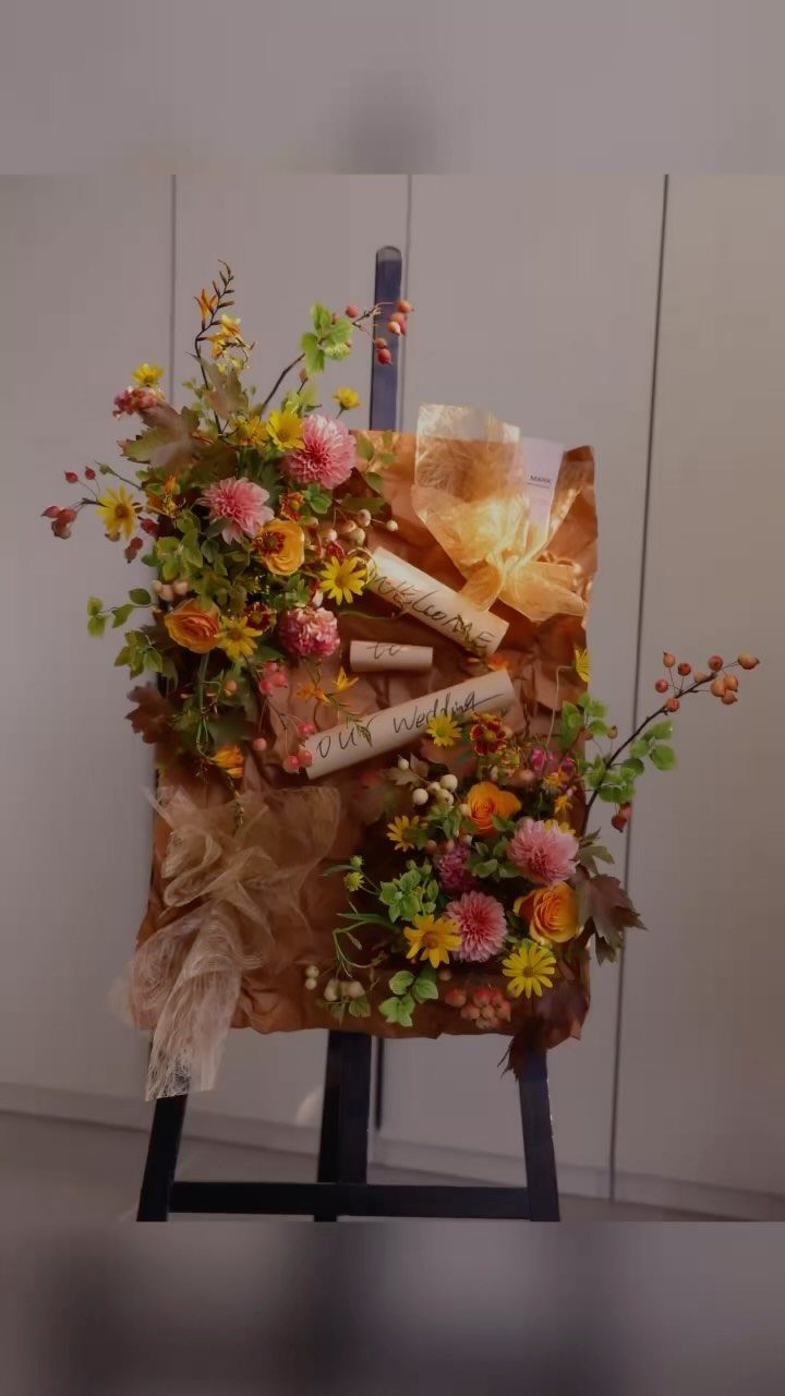 28 Wedding Decorations Ideas at Home with Flowers and Lights 26 Floral arrangements and parchment scrolls on a brown backdrop supported by a black easel.