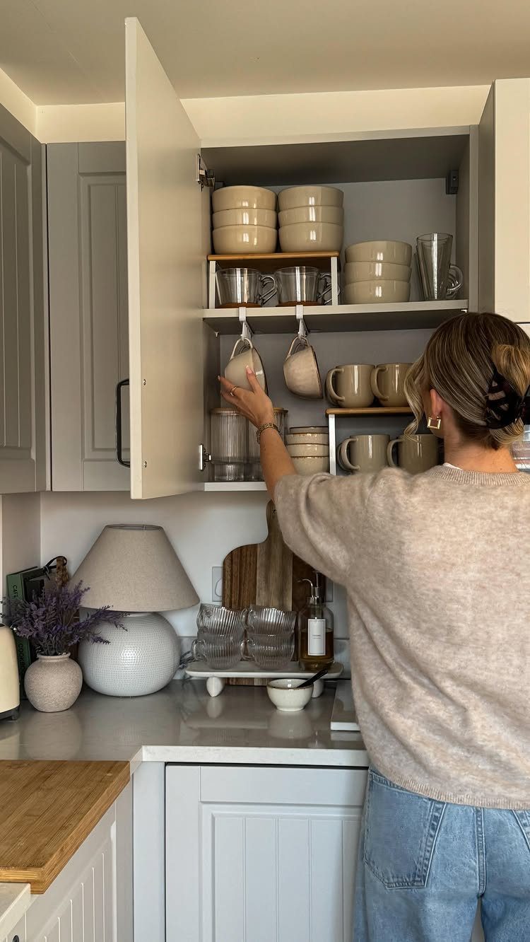 Woman reaching for glasses stored on white shelf risers inside a kitchen cabinet.