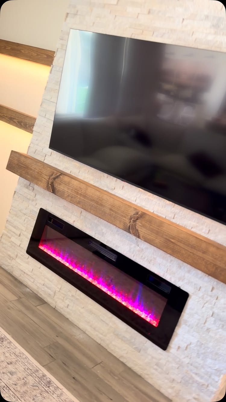 16 TV Designs for Living Room Walls, Units and Mounts 5 Television mounted on a white stone wall above a wooden mantel and electric fireplace.