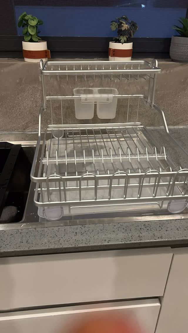 Silver metal two-tier dish drying rack on a grey kitchen countertop near a sink.