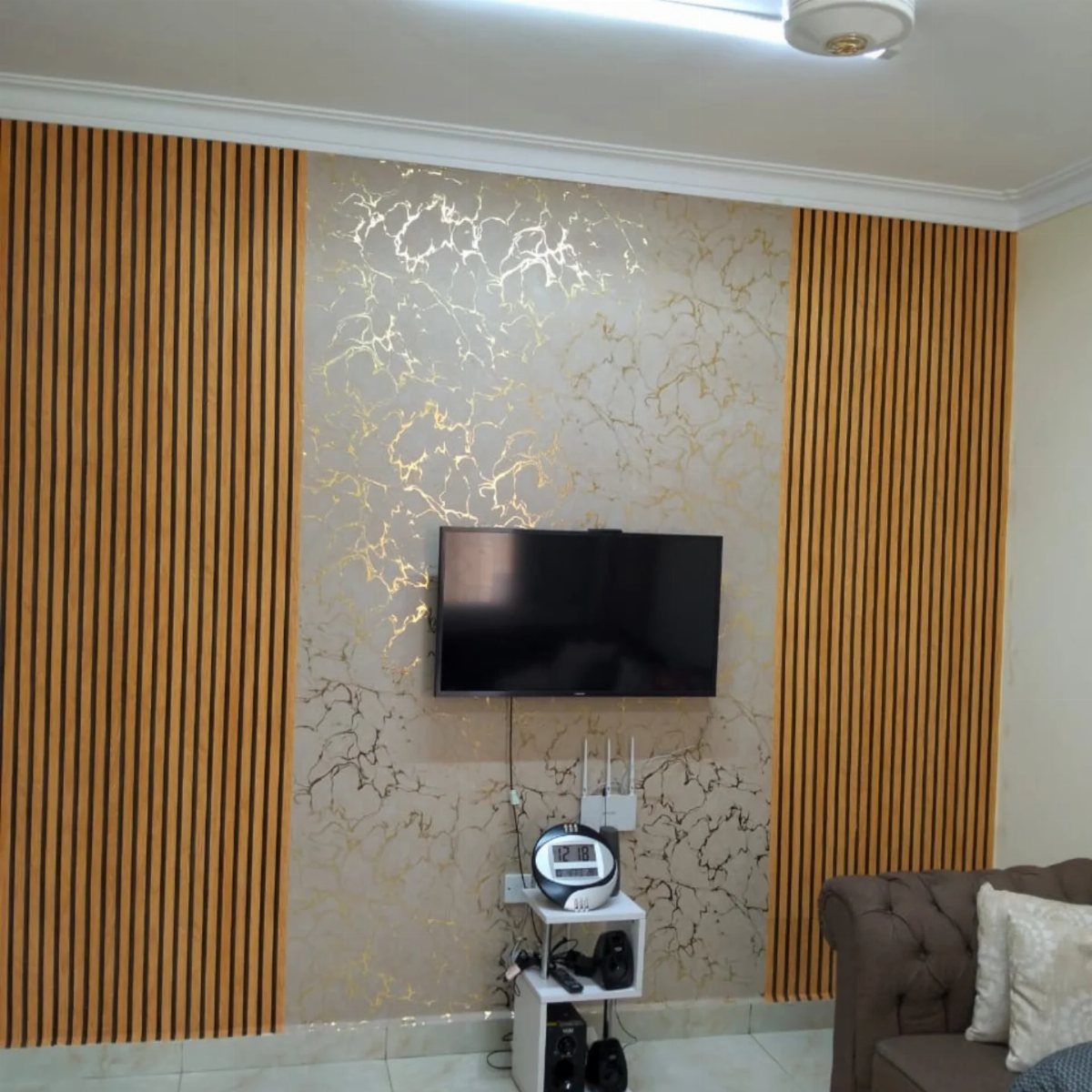 16 TV Designs for Living Room Walls, Units and Mounts 6 Wall-mounted television on gold marble wallpaper flanked by vertical light-wood slat panels.