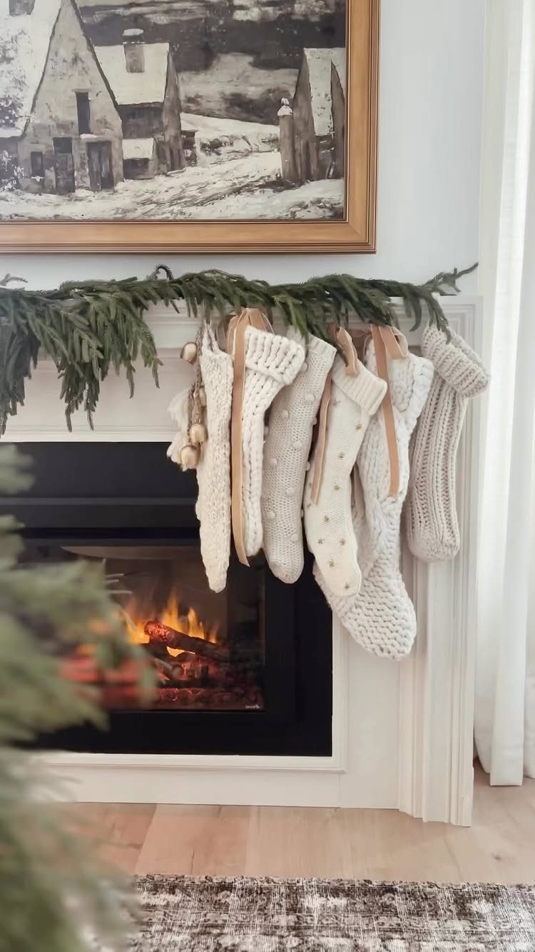 White fireplace mantel with green pine garland and cream knit stockings above a fire.